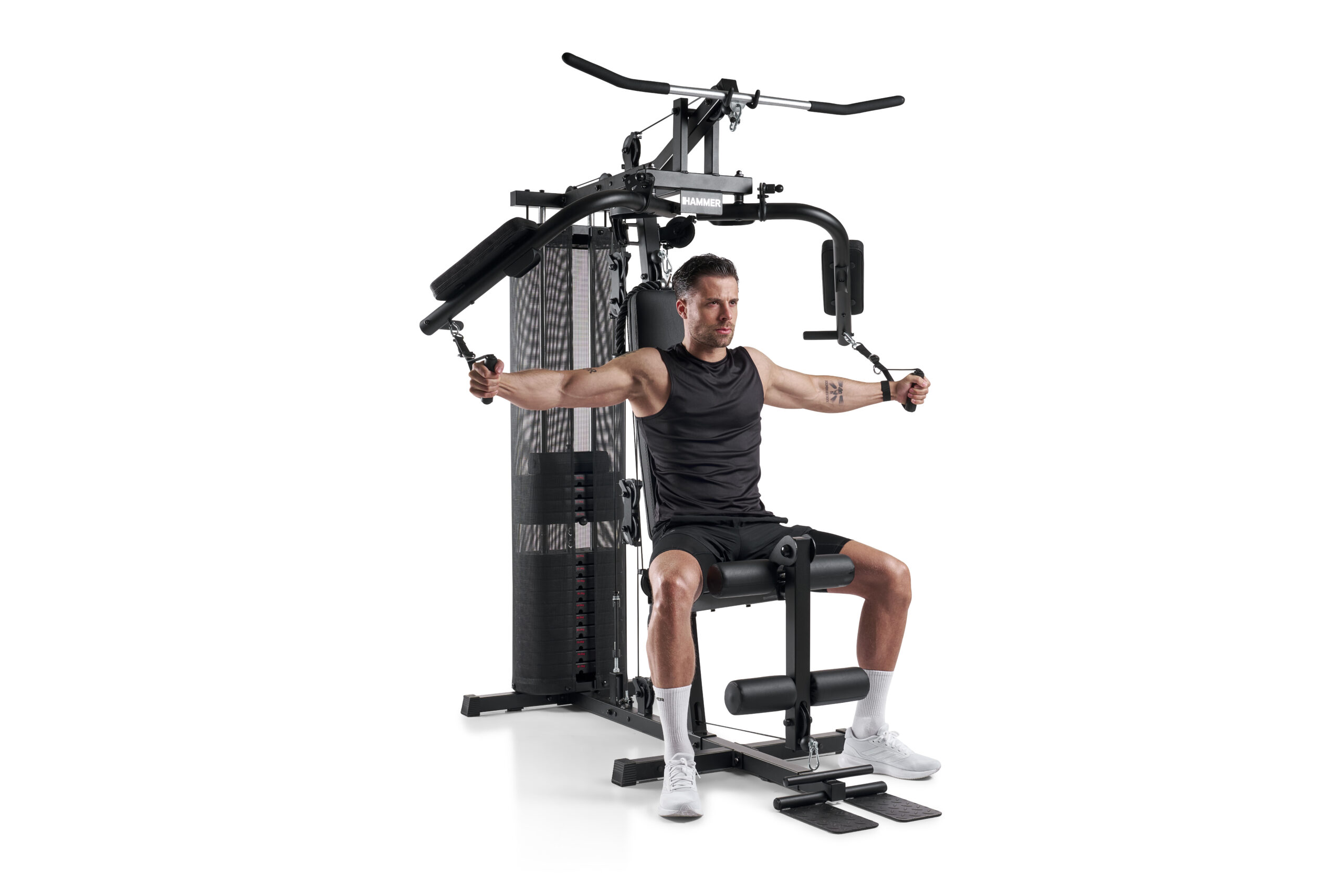 Man in a sleeveless shirt sits in a multi-station gym machine, pressing outward on cables with arms extended in a chest press motion.