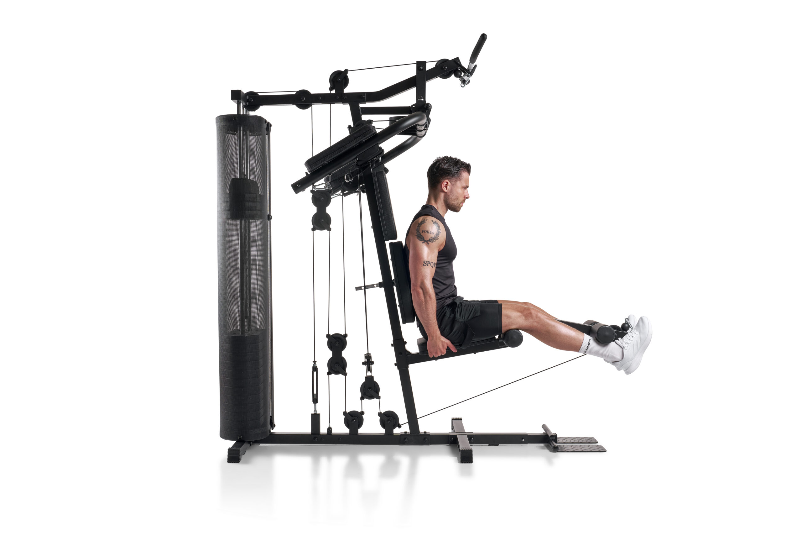 Man seated on a multi-gym machine performing leg extension with arms gripping handles and ropes overhead.