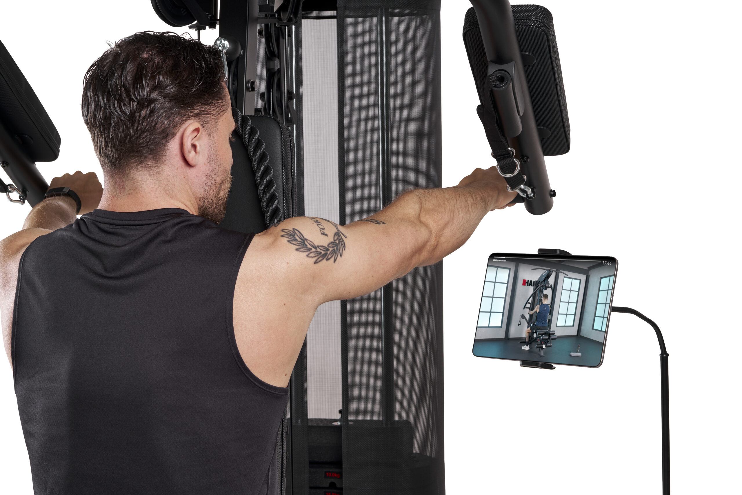 Man using a multi-gym, gripping handles while a tablet shows a gym workout on a stand nearby