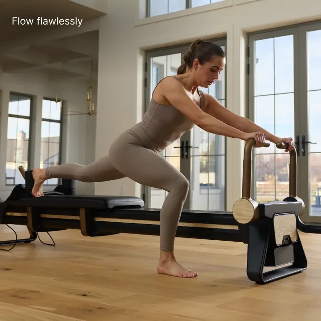 Woman in beige athletic wear performs a Pilates lunge on a reformer in a bright studio with large windows.