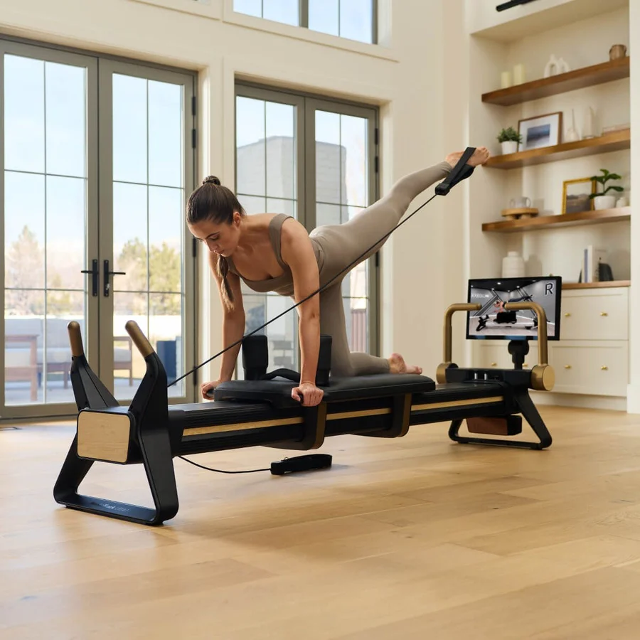 Woman in beige workout attire on a Pilates reformer, balancing on hands and knees while pulling a strap for resistance in a bright home studio with large windows.