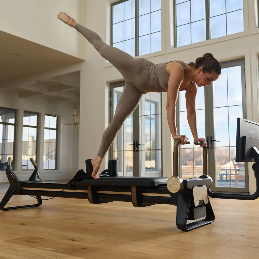 Woman in beige activewear balances on a Pilates reformer, one leg extended behind and hands gripping the handles in a bright studio.