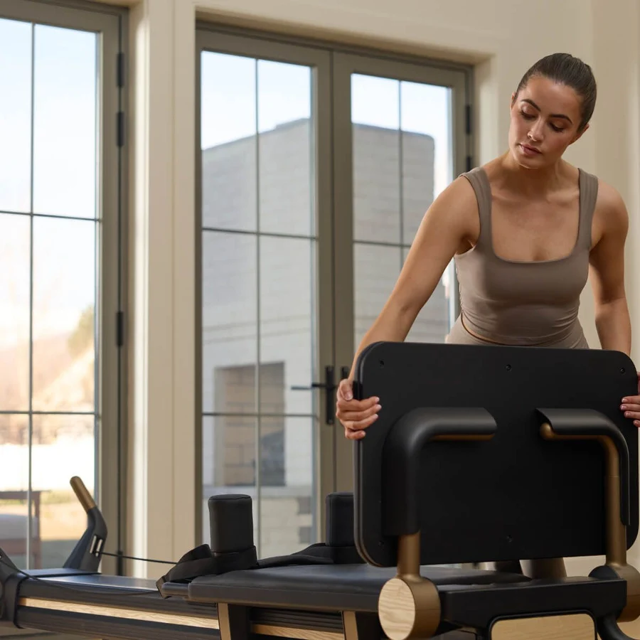 Woman in a gray tank top adjusts a Pilates reformer in a bright studio windowed room.