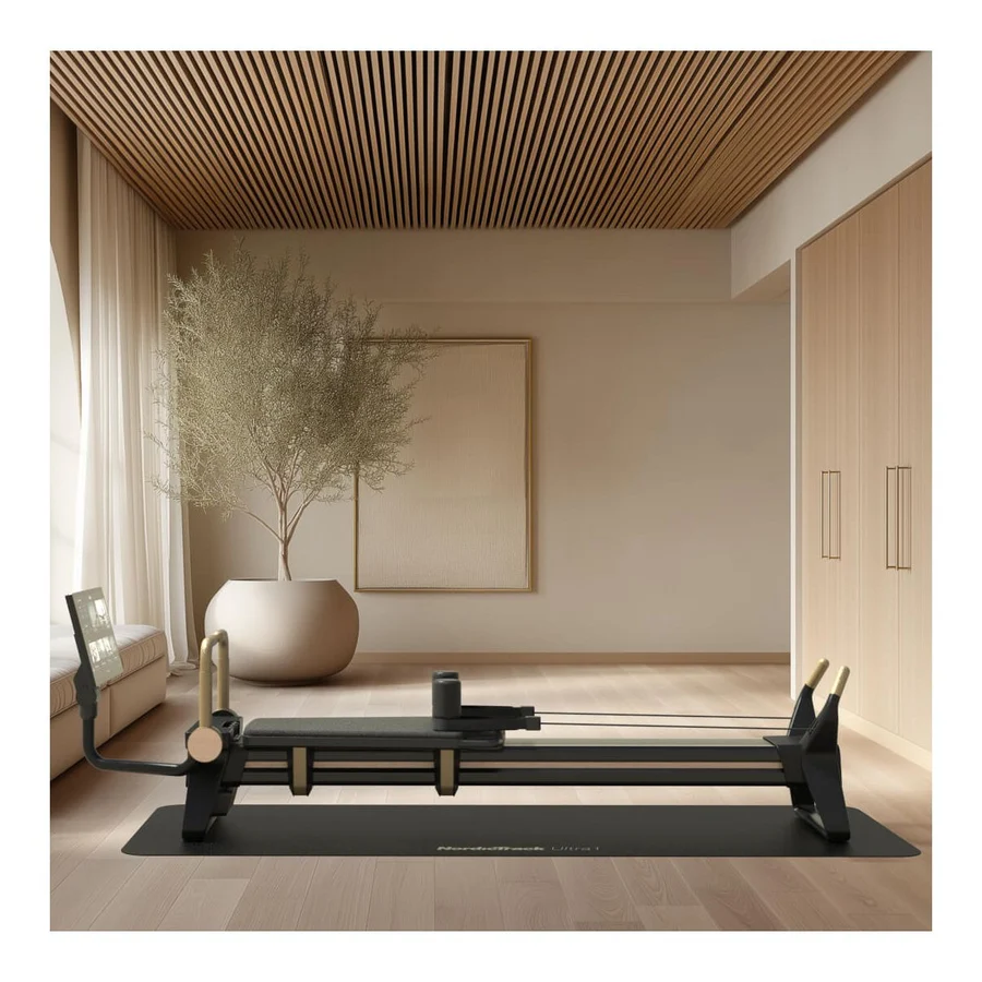 A modern home gym with a black Pilates reformer on a mat, in a bright room with wooden floors and a large potted plant in the background.
