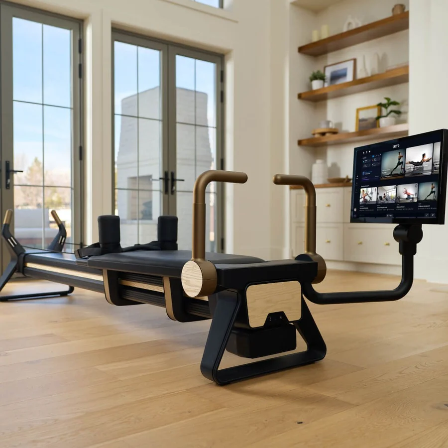High-end Pilates reformer with a mounted screen in a bright home gym, featuring tan handles and wooden accents on a light wood floor.