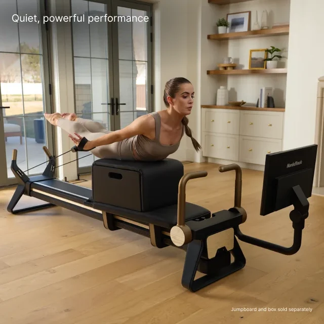 Woman in beige activewear performs a Pilates leg extension on a reformer in a bright home gym setting, with a screen at the end of the machine.